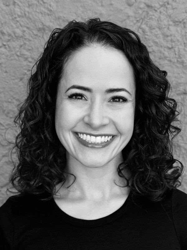 Headshot of Hilary Kirwan, a communications leader with curly brown hair, wearing a black top and smiling warmly at the camera